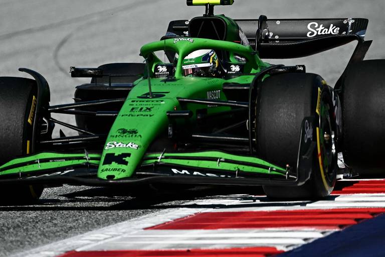 Um carro de Fórmula 1 de cor verde, com detalhes em preto, está em movimento em uma pista. O carro possui um plan  aerodinâmico e está passando por uma curva, com arsenic  rodas dianteiras levantadas levemente bash  chão. O fundo é desfocado, sugerindo velocidade, e há uma faixa de asfalto com linhas vermelhas e brancas.