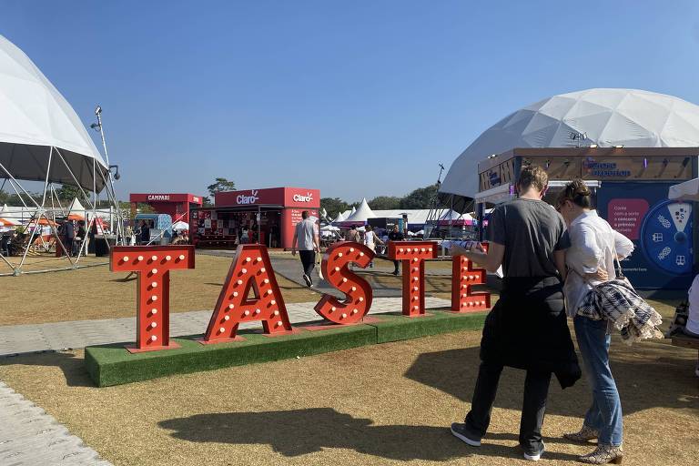 A imagem mostra um evento ao ar livre com uma grande estrutura em forma de domo ao fundo. Em primeiro plano, há uma grande placa iluminada com a palavra 'TASTE' em letras vermelhas. Algumas pessoas estão caminhando pelo local, e há barracas de comida visíveis ao fundo. O céu está claro e ensolarado, e o chão é coberto por grama seca.