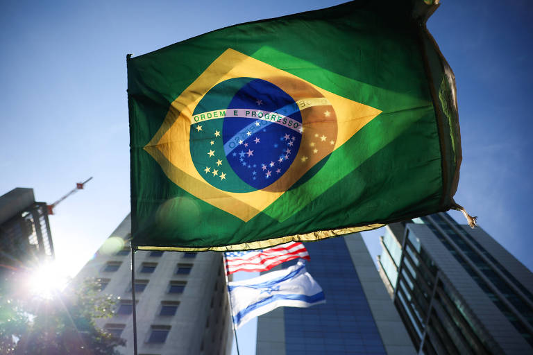 A imagem mostra a bandeira do Brasil tremulando ao vento, com o sol brilhando ao fundo. A bandeira é verde com um losango amarelo e um círculo azul, contendo estrelas brancas. Ao fundo, é possível ver edifícios modernos e outra bandeira, possivelmente da cidade de São Paulo, em cores azul e branca.