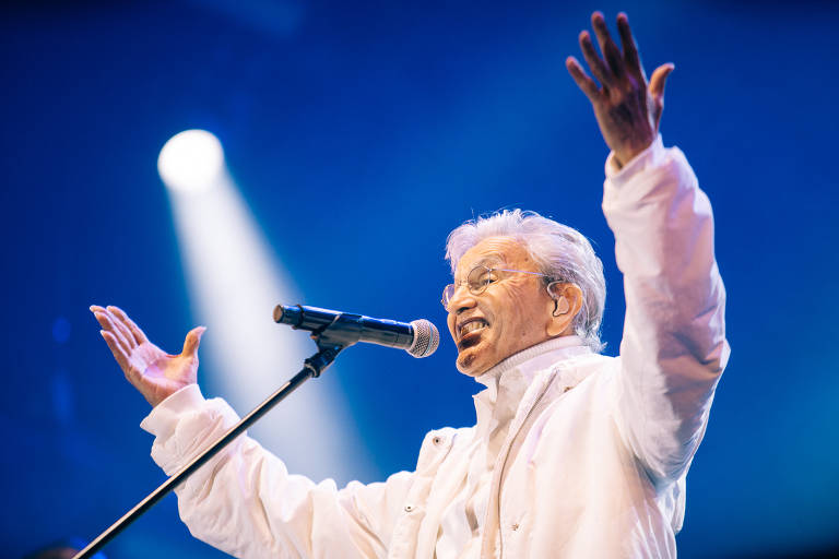 Artist performing on stage wearing a white costume. He stands with his arms raised and a microphone in his hand, smiling as he sings. The background is illuminated with blue and white lights, creating a vibrant atmosphere.