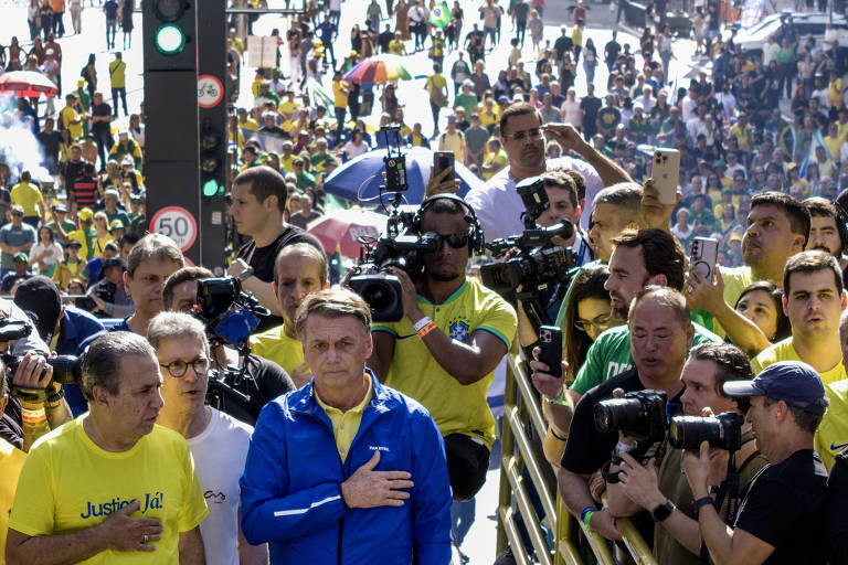 A imagem mostra uma grande multidão em uma manifestação, com muitas pessoas vestindo camisetas amarelas. No centro, um homem de jaqueta azul está com a mão sobre o coração, cercado por câmeras e repórteres. Ao fundo, há uma grande quantidade de pessoas, algumas segurando bandeiras e cartazes. O ambiente parece ser de um evento ao ar livre, com um clima de celebração e apoio.