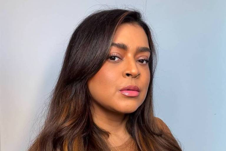 A imagem mostra uma mulher com cabelo longo e ondulado, usando uma blusa marrom. Ela está olhando para a câmera com uma expressão séria. O fundo é de uma parede clara, e a iluminação é suave, destacando os detalhes do cabelo e do rosto.