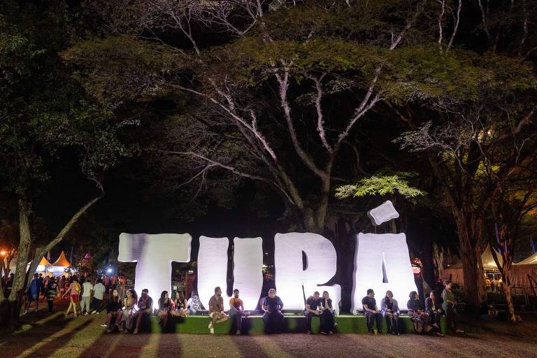 A imagem mostra um grande letreiro iluminado com a palavra 'TUBÁ' em letras brancas, situado em um espaço ao ar livre à noite. Várias pessoas estão sentadas e em pé ao redor do letreiro, que está cercado por árvores e um ambiente festivo. Ao fundo, há tendas e outras pessoas, sugerindo um evento ou celebração.