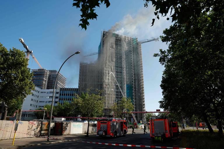 A imagem mostra um edifício em construção com fumaça saindo do topo. Há guindastes ao fundo e várias árvores na frente. Veículos de bombeiros estão estacionados na área, e há uma barreira de segurança visível. O céu está claro, indicando um dia ensolarado.