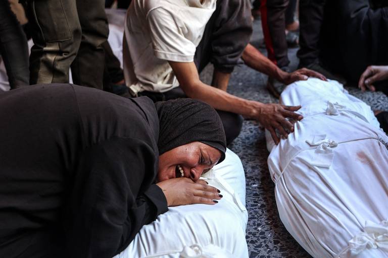 De afbeelding toont verschillende mensen in een rouwende omgeving, met de focus op een vrouw met een zwarte hijab, die op een met een wit laken bedekt lichaam ligt en pijn en verdriet uitdrukt. Op de achtergrond zijn andere mensen in de buurt, sommigen raken de lichamen aan, terwijl anderen de scène met een verdrietige blik bekijken.