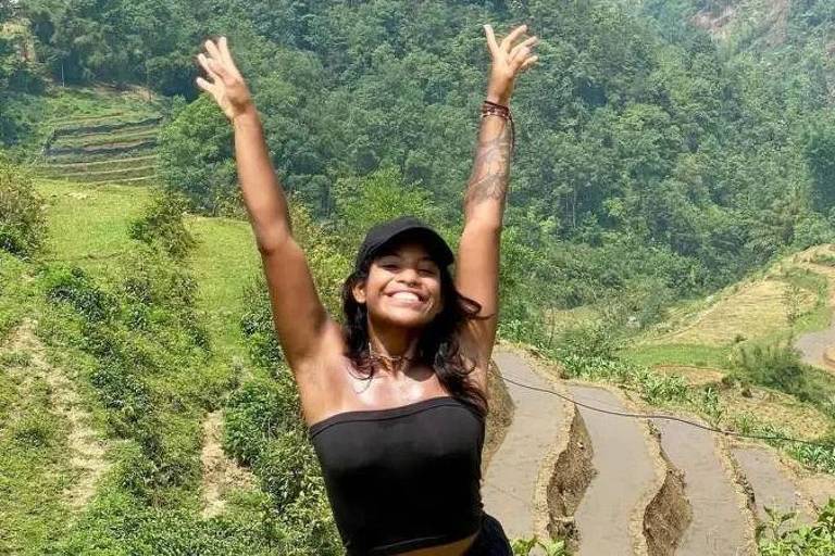 A woman stands in a mountainous landscape, her hands raised in joy. She is wearing a black blouse and a cap. In the background are terraced rice fields and lush green vegetation, with mountains in the distance.