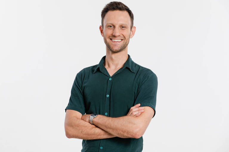 Um homem está posando para a foto com um sorriso. Ele tem cabelo curto e usa uma camisa verde escura. Os braços estão cruzados sobre o peito e o fundo da imagem é branco.