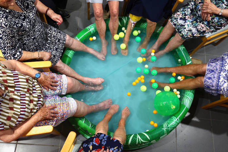 A imagem mostra várias pessoas sentadas ao redor de uma piscina inflável com água azul. Os pés de várias pessoas estão imersos na água, e há bolinhas coloridas flutuando na superfície. As pessoas estão vestindo roupas de verão, e a cena transmite um ambiente descontraído e divertido.