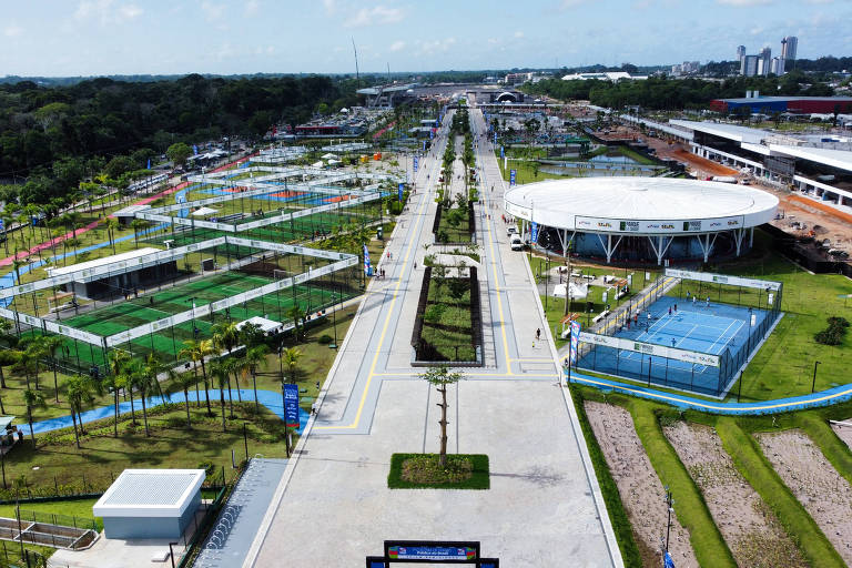 A imagem mostra uma vista aérea de um parque esportivo, com várias quadras e campos visíveis. Há uma grande área pavimentada que se estende ao longo do parque, cercada por árvores e áreas verdes. No fundo, pode-se ver um edifício e uma área em construção. O céu está claro e ensolarado, e a disposição das quadras sugere um espaço dedicado a atividades esportivas e recreativas.