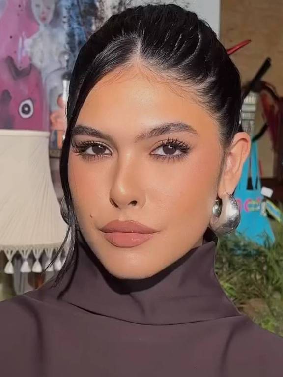 A imagem mostra uma mulher com cabelo preso em um coque, usando uma blusa de gola alta na cor marrom. Ela tem maquiagem bem feita, com destaque para os olhos e lábios. Ao fundo, há elementos decorativos, incluindo uma lâmpada e objetos coloridos.