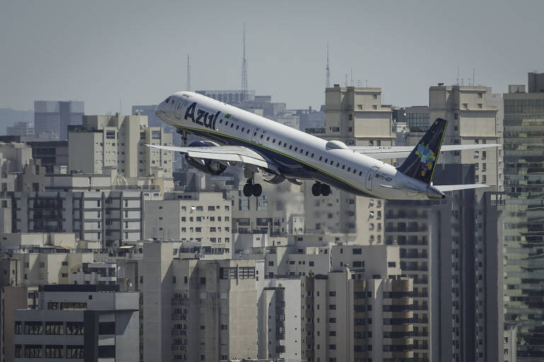 Um avião da companhia aérea Azul está decolando em direção ao céu, com a cidade ao fundo. O avião é branco com detalhes em azul e verde, e está em um ângulo ascendente, enquanto vários prédios altos e modernos compõem a paisagem urbana ao redor.