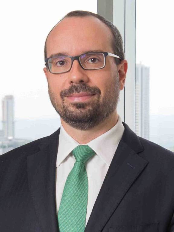 Um homem está posando para a foto em um ambiente de escritório. Ele usa um terno escuro, uma camisa branca e uma gravata verde. O homem tem cabelo curto e usa óculos. Ao fundo, há janelas que mostram uma vista urbana com prédios.