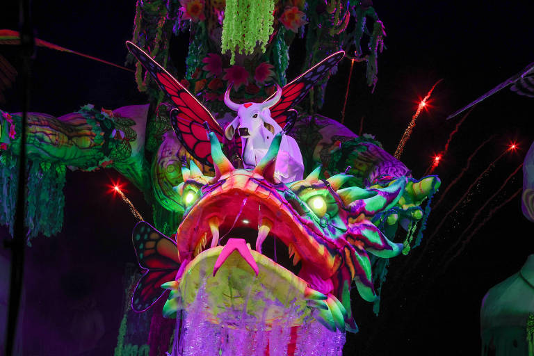 A imagem mostra uma grande figura de um dragão colorido, com detalhes vibrantes em verde, rosa e amarelo. O dragão possui uma cabeça grande com dentes afiados e olhos brilhantes. Em cima da cabeça do dragão, há uma figura de um boi branco, e ao fundo, há elementos decorativos como flores e luzes. O ambiente é festivo, com fogos de artifício visíveis ao redor, criando uma atmosfera de celebração.