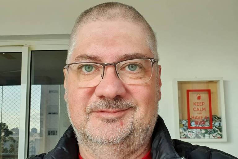 Um homem com cabelo curto e grisalho, usando óculos e uma jaqueta preta. Ele está sorrindo e posando para a câmera. Ao fundo, há uma janela com vista para a cidade e uma parede com um quadro que diz 'KEEP CALM'.