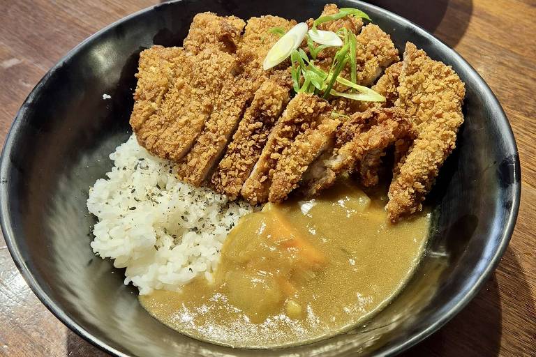 A imagem mostra um prato de comida japonesa com arroz branco, curry e pedaços de carne empanada e frita, dispostos de forma organizada. Ao fundo, há outro prato com frango frito e salada, além de uma porção de cebola roxa em conserva. O ambiente parece ser um restaurante, com mesas e cadeiras visíveis.