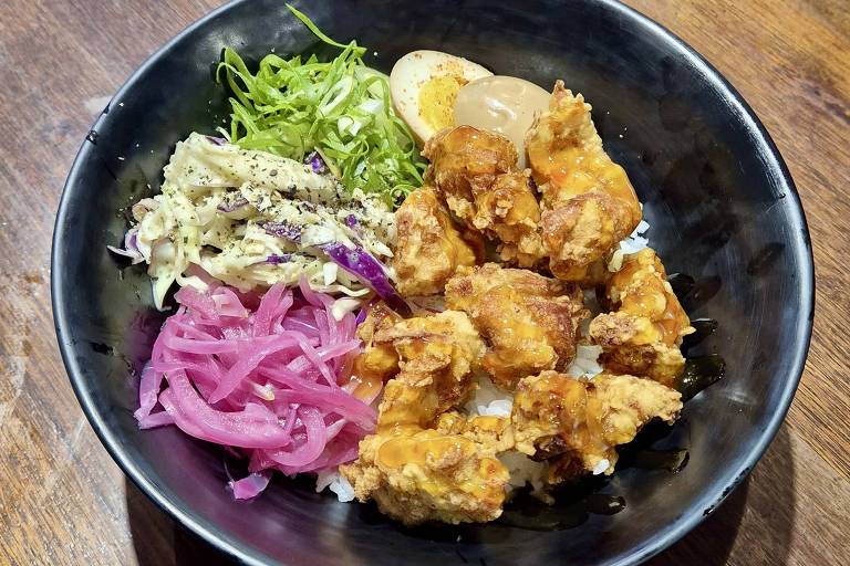 Um prato de comida japonesa apresenta uma porção de arroz coberta com pedaços de frango frito, acompanhada de vegetais como repolho roxo, cebola roxa em conserva, alface e um ovo cozido. O prato é servido em uma tigela preta sobre uma mesa de madeira.
