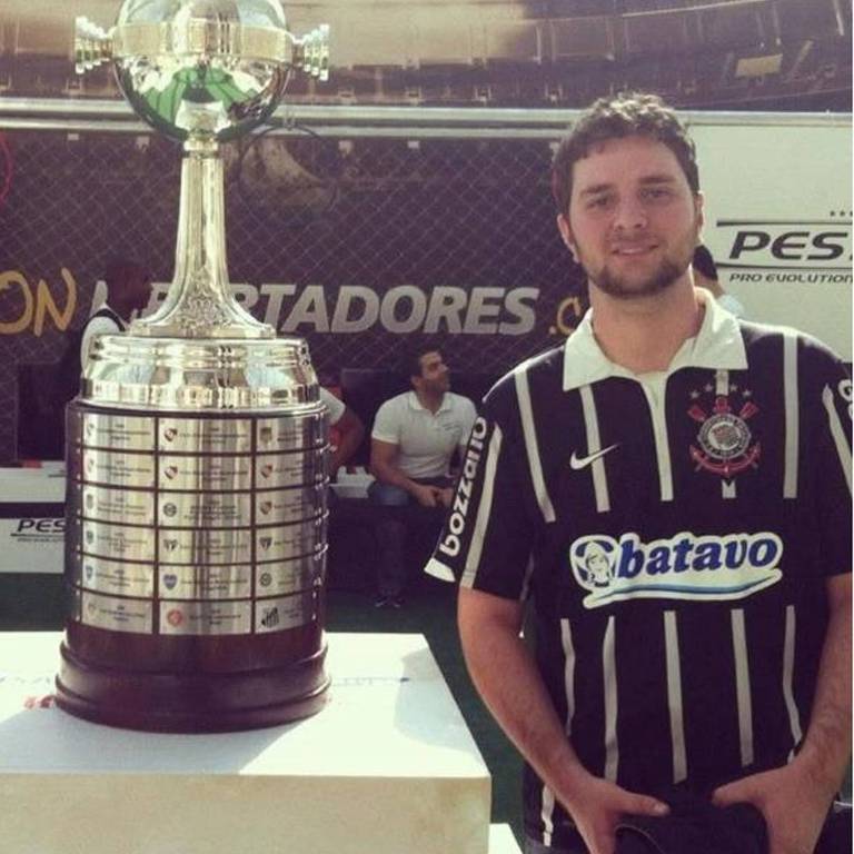 A man is posing next to a Copa Libertadores trophy. He is wearing a black and white striped shirt. The trophy is large and shiny, with a wide base and a sphere on top. In the background, there is a decoration that refers to the Copa Libertadores event.