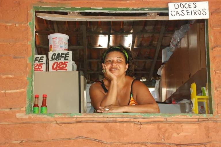 A imagem mostra uma mulher sorridente, com cabelo preso e usando uma blusa de alças, apoiada em uma janela. Ela está em um ambiente rústico, com paredes de barro e um teto de madeira. Ao fundo, há prateleiras com produtos, incluindo caixas de café e um recipiente de doces. Acima da janela, há uma placa escrita 'DOCES CASEIROS'.