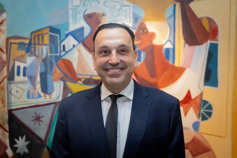 Um homem sorridente est posicionado em primeiro plano, vestindo um terno escuro e uma gravata. Ao fundo, h um mural colorido que apresenta figuras humanas estilizadas e elementos arquitetnicos. As cores do mural incluem tons de azul, laranja e amarelo, criando um contraste vibrante com a roupa do homem.