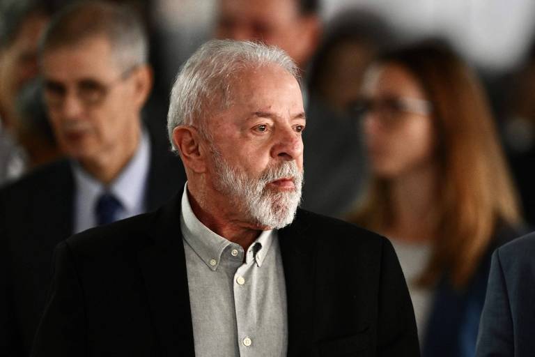 A imagem mostra um grupo de pessoas em um evento. Em primeiro plano, um homem de cabelo grisalho e barba branca, vestindo uma camisa clara e um paletó escuro, parece pensativo. Ao fundo, outras pessoas estão visíveis, algumas com expressões neutras, e outras em conversação. A iluminação é suave e o ambiente parece ser interno.