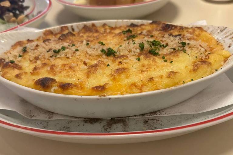 A imagem mostra um prato de gratinado, com uma crosta dourada e crocante, polvilhado com ervas verdes. O prato é oval e está sobre uma bandeja. Ao fundo, pode-se ver outros pratos, incluindo um com bacon e outro com um tipo de sobremesa. A mesa é clara e o ambiente parece ser de um restaurante.