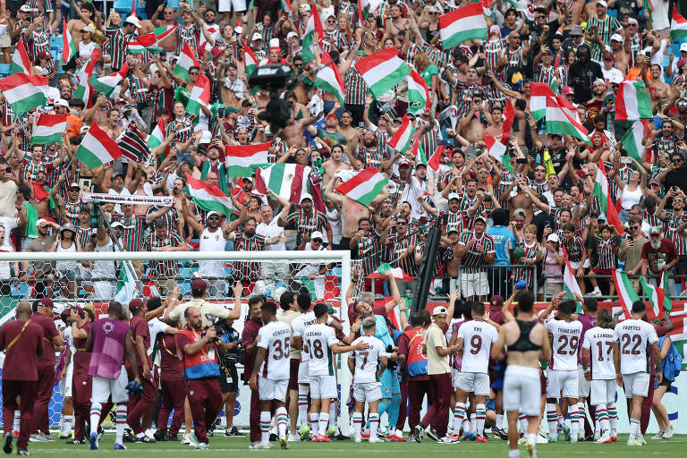 A imagem mostra uma grande quantidade de torcedores do Fluminense em um estádio, todos vestindo camisas listradas em verde, vermelho e branco. Eles estão segurando bandeiras nas mesmas cores e demonstrando entusiasmo. No primeiro plano, alguns jogadores da equipe estão se reunindo, enquanto a torcida celebra ao fundo.