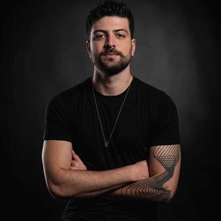 Um homem está posando para a foto com os braços cruzados. Ele usa uma camiseta preta e um colar prateado. O fundo é escuro, criando um contraste com sua pele clara e cabelo escuro. O homem tem uma tatuagem geométrica no braço direito.