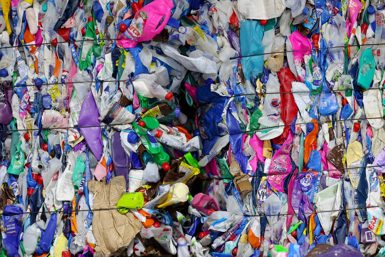 A imagem mostra uma grande pilha de plásticos recicláveis, incluindo garrafas e sacolas de diversas cores, como azul, verde, rosa, roxo e branco. Os plásticos estão organizados em uma estrutura de malha, evidenciando a variedade de tipos e cores dos materiais recicláveis.