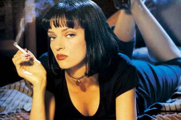 A imagem mostra uma mulher de cabelo preto e liso, deitada em uma cama. Ela está usando uma blusa preta e tem uma expressão séria. A mulher segura um cigarro com a mão esquerda, enquanto a mão direita está apoiada na cama. Ao fundo, é possível ver uma perna de outra pessoa, sugerindo que há alguém mais na cena. A iluminação é suave, criando um ambiente intimista.