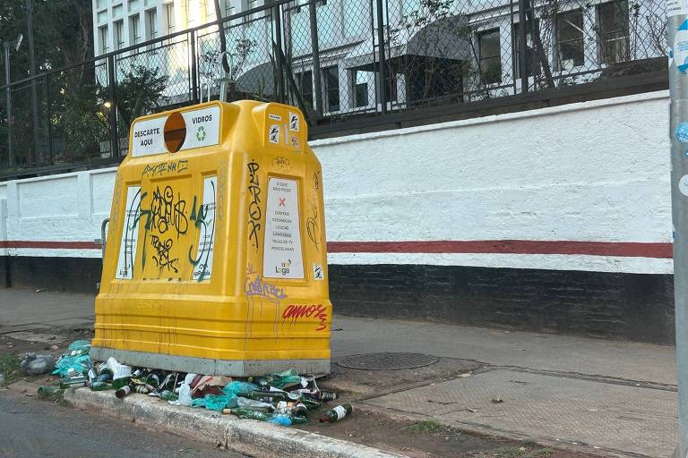 De afbeelding toont een gele prullenbak op een stoep, omringd door opgehoopt afval, waaronder flessen. De prullenbak is beschilderd met tekst en graffiti. Op de achtergrond is een witte muur met een rode streep en een hek te zien, wat een stedelijke omgeving suggereert. De scène lijkt een drukke straat te zijn, met de prullenbak centraal in de afbeelding.