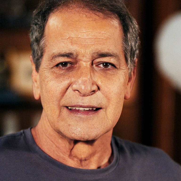 Um homem de pele clara e cabelo grisalho, com um sorriso leve, está olhando para a câmera. Ele usa uma camiseta cinza e está em um ambiente interno, com um fundo desfocado que sugere uma sala de estar. A iluminação é suave, destacando suas características faciais.