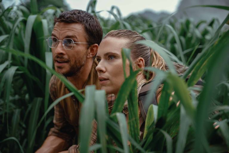Um homem e uma mulher estão agachados entre altas plantas verdes, parecendo atentos e curiosos. O homem usa óculos e tem cabelo curto, enquanto a mulher tem cabelo preso em um coque. Ambos estão vestidos de forma casual e observam algo à frente com expressões de concentração.