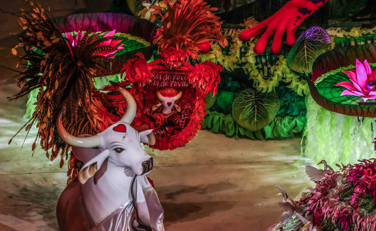 A imagem mostra uma decoração vibrante de festa, com destaque para um boi branco estilizado, adornado com um lenço prateado. Ao fundo, há arranjos de plantas e flores coloridas, incluindo folhas verdes e flores rosa. Elementos como penas e outros adornos naturais estão presentes, criando um ambiente festivo e exuberante.