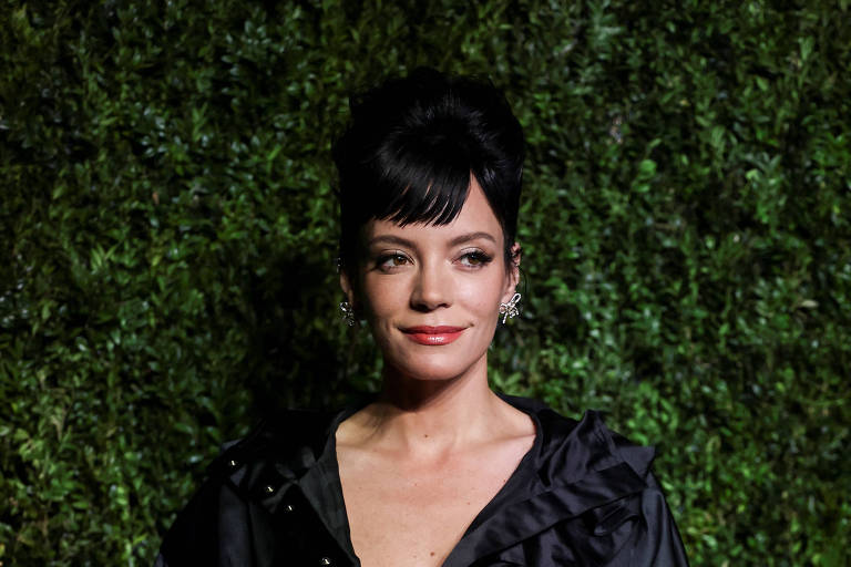 A imagem mostra uma mulher em um evento, vestindo uma jaqueta preta com detalhes em ruffles. Ela tem cabelo preto e liso, preso em um coque, e está posando em frente a um fundo verde. A expressão dela é sorridente e confiante.