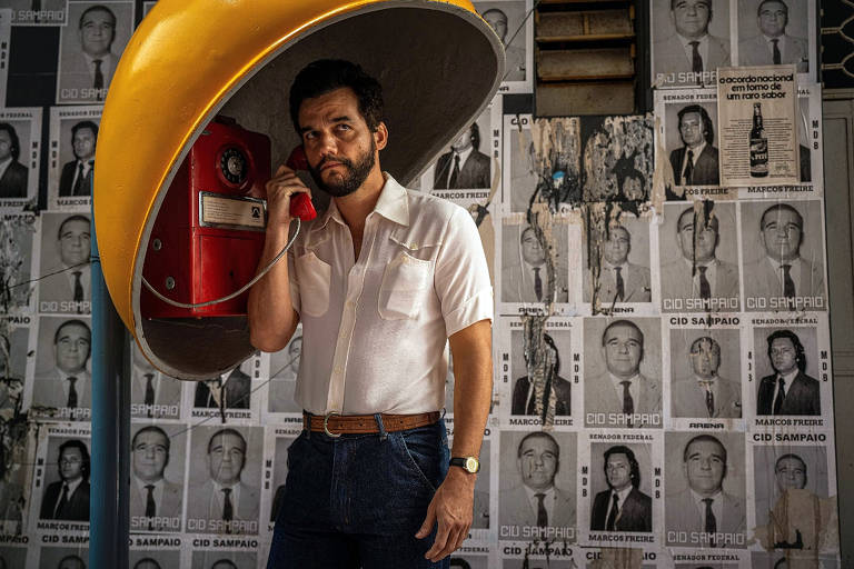 Um homem está em pé, segurando um telefone vermelho em um ambiente decorado com várias fotos em preto e branco de pessoas. Ele usa uma camisa branca e jeans escuros, e está posicionado sob uma estrutura amarela em forma de cúpula. O fundo é repleto de cartazes com rostos e nomes, sugerindo um tema de nostalgia ou história.