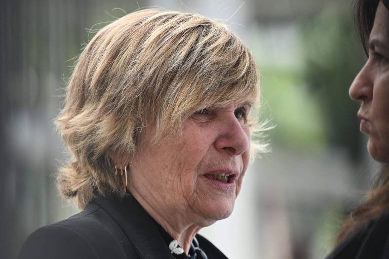 A imagem mostra duas mulheres em um ambiente externo. A mulher à esquerda tem cabelo loiro e curto, usando uma blusa preta e um colar. Ela parece estar falando e tem uma expressão facial atenta. A mulher à direita tem cabelo escuro e longo, e está olhando para a mulher à esquerda, com uma expressão séria. O fundo é desfocado, sugerindo que a cena ocorre em um local público.