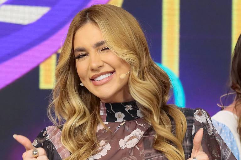 Uma mulher com cabelo longo e ondulado, vestindo uma blusa transparente com estampas florais e uma saia preta. Ela está sorrindo e fazendo sinal de positivo com ambas as mãos. Ao fundo, há um cenário colorido e moderno, típico de um programa de televisão.