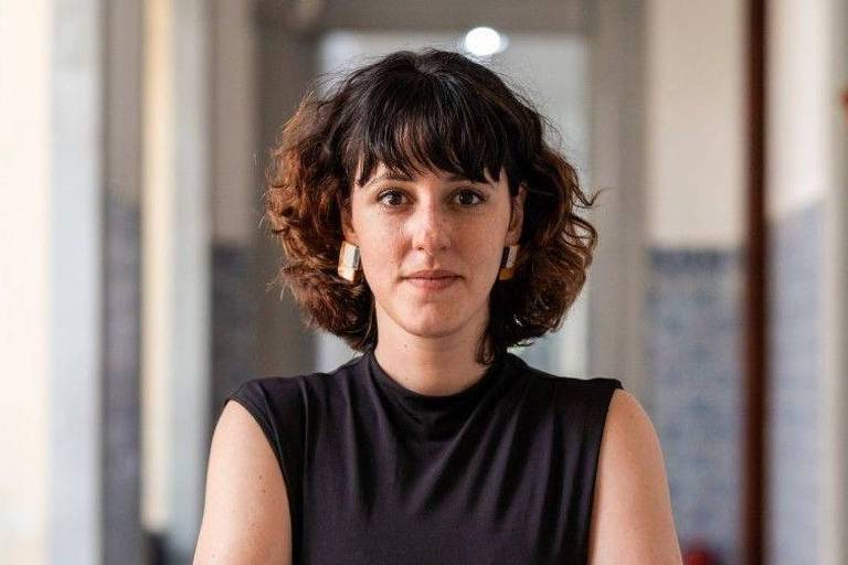 A imagem mostra uma mulher com cabelo cacheado e escuro, usando uma blusa preta. Ela está em um ambiente interno, com paredes claras e um piso que parece ser de azulejos. A mulher está olhando diretamente para a câmera, com uma expressão neutra. Ao fundo, há uma iluminação suave e um corredor visível.