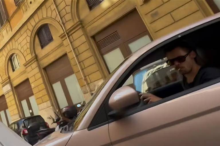A imagem mostra um homem sentado no banco do motorista de um carro. Ele usa óculos escuros e parece estar prestes a dirigir. O carro é de cor rosa e está estacionado em uma rua com prédios de estilo arquitetônico clássico ao fundo. É possível ver algumas motos estacionadas ao lado do carro.