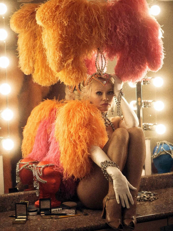 A imagem mostra uma artista de palco sentada em um camarim, cercada por luzes brilhantes. Ela usa um traje com plumas coloridas em tons de rosa e laranja, e está com uma pose elegante, segurando a cabeça com uma das mãos. O ambiente é decorado com espelhos e produtos de maquiagem dispostos em uma superfície próxima.