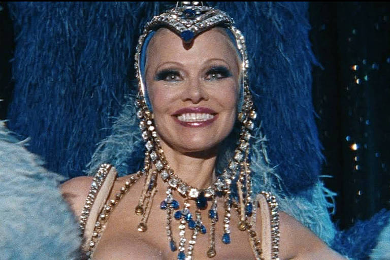 Uma mulher sorridente está em uma apresentação, usando um traje brilhante e elaborado, adornado com joias e penas. O fundo é escuro, com luzes que destacam a figura dela, criando um ambiente de espetáculo.