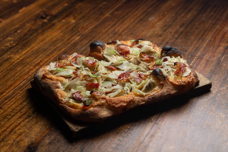 A imagem mostra uma pizza retangular com uma crosta dourada e levemente queimada nas bordas. A cobertura é composta por cebolas fatiadas, pedaços de linguiça e ervas, dispostos de maneira uniforme sobre a massa. A pizza está sobre uma tábua de madeira, com um fundo de mesa também de madeira, criando um ambiente rústico.