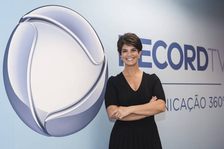 Uma mulher com cabelo curto e escuro está sorrindo e posando ao lado do logotipo da RecordTV, que é uma esfera estilizada. Ela está vestindo uma blusa preta e está em um ambiente com uma parede azul clara. Ao lado do logotipo, está escrito 'RECORDTV' e 'COMUNICAÇÃO 360°'.