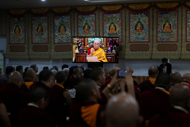 A imagem mostra uma sala cheia de monges budistas, todos vestidos com túnicas marrom e laranja. No fundo, há uma tela grande exibindo a imagem de um líder espiritual, que está falando e segurando um papel. As paredes estão decoradas com tapeçarias que retratam figuras budistas. Alguns monges estão olhando para a tela, enquanto outros estão em conversas ou tirando fotos.