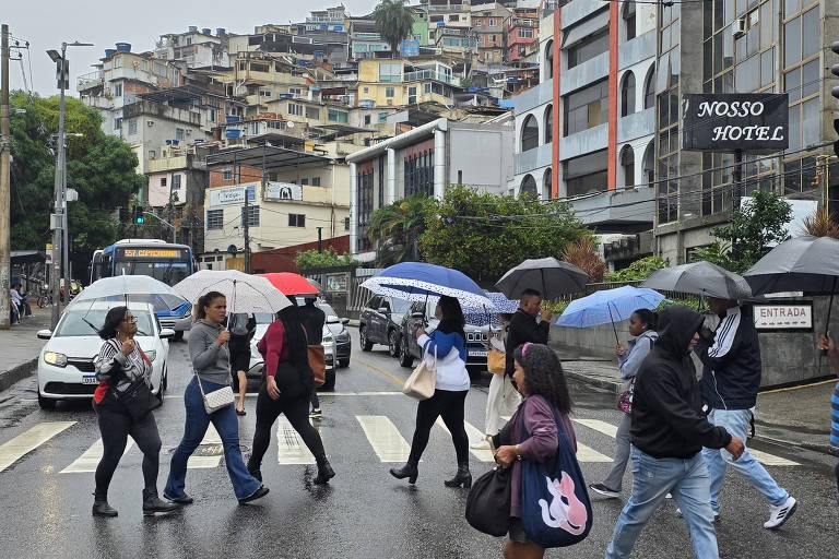 A imagem mostra um grupo de pessoas atravessando uma rua em um dia chuvoso, segurando guarda-chuvas. Ao fundo, há um morro com casas empilhadas e um ônibus na rua. À direita, um edifício com a placa "NOSSO HOTEL" é visível. O asfalto está molhado devido à chuva.