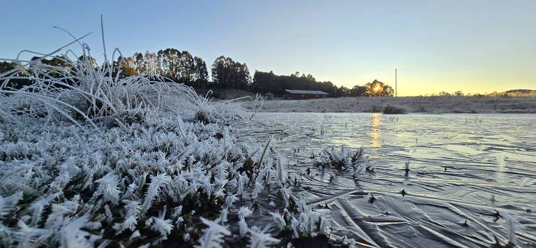 A imagem mostra uma paisagem ao amanhecer, com grama coberta de geada branca e um lago parcialmente congelado. Ao fundo, há árvores e uma construção iluminada pelo sol nascente. O céu apresenta tons de azul e laranja, indicando o início do dia.