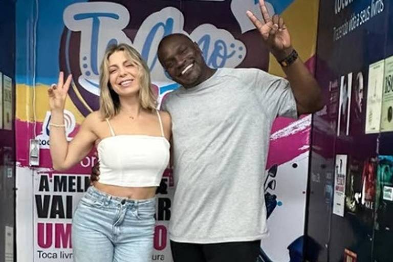 Duas pessoas estão posando para a foto em um ambiente interno. A mulher, com cabelo loiro e liso, usa uma blusa branca e calças jeans largas. O homem, com pele escura, está vestido com uma camiseta cinza e calças pretas. Ambos fazem o sinal de paz com as mãos. Ao fundo, há uma parede decorada com cores vibrantes e textos relacionados a um concurso cultural.
