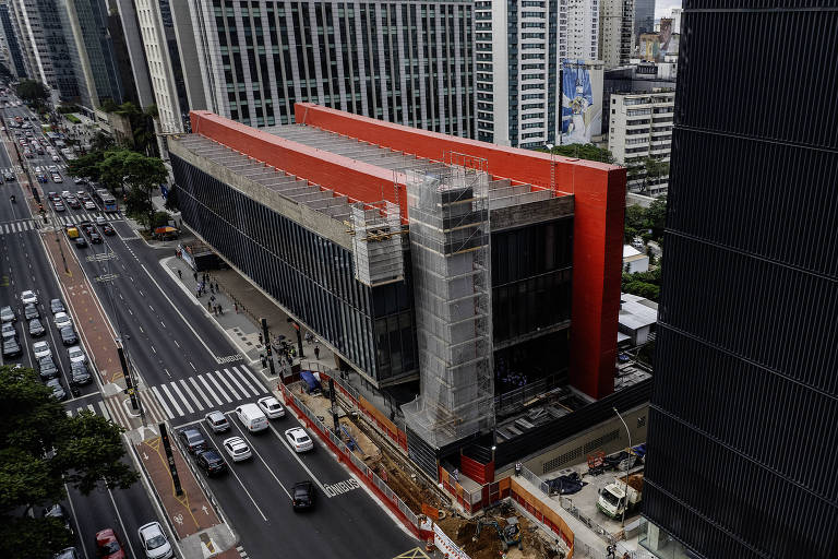 A imagem mostra uma área urbana com edifícios altos. À esquerda, há um edifício em construção com uma fachada vermelha e partes cobertas por uma estrutura de proteção. À direita, um edifício de fachada escura. A rua em frente tem tráfego de veículos e há uma obra em andamento na calçada, com cercas de proteção ao redor. O céu está nublado.