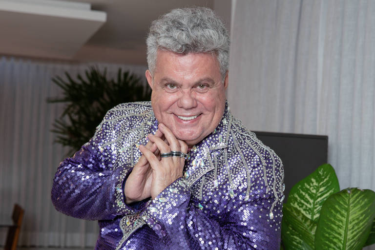 Um homem sorridente está posando em um ambiente interno. Ele usa um traje brilhante de cor roxa com lantejoulas. O fundo apresenta cortinas brancas e uma planta verde ao lado. O homem tem cabelo grisalho e está com as mãos juntas em uma pose amigável.
