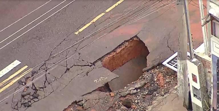 A imagem mostra um buraco grande na rua, com as bordas irregulares e rachaduras visíveis ao redor. O asfalto está danificado, e há uma parte exposta do solo abaixo. Ao lado do buraco, há uma calçada e um poste de luz.